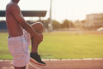 Midsection of shirtless athlete stretching leg on sports track