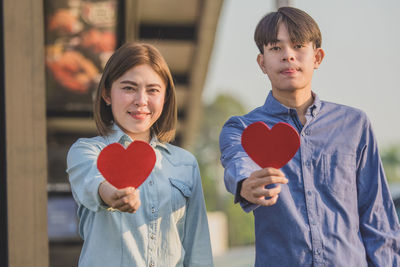 Portrait of smiling couple holding heart shape while standing outdoors
