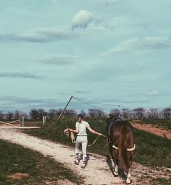 Rear view of woman walking with brown horse on footpath against sky