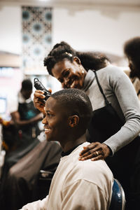Smiling female barber cutting hair of male customer with electric razor at salon