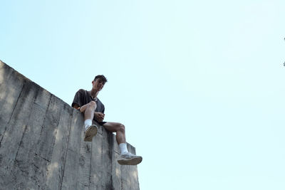 From below of smiling male sitting on high wall of concrete building and relaxing during parkour training in city