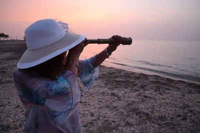 Woman looking through hand-held telescope at beach against sky during sunset