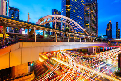 Light trails on bridge by buildings against sky at night