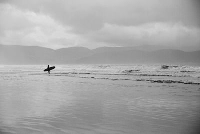 Distant view of surfer with surfboard on shore against mountains