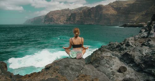 Rear view of woman sitting on rock by sea against mountain