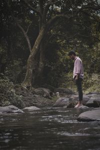 Side view of young man standing by river against trees in forest