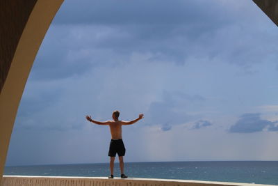 Rear view of shirtless man with arms outstretched standing on retaining wall by sea against sky