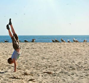 Side view of boy doing handstand at beach against sky
