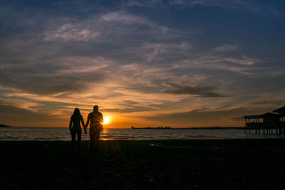 Silhouette couple walking on shore at beach against sky during sunset