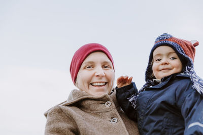 Portrait of smiling woman carrying daughter against sky during winter