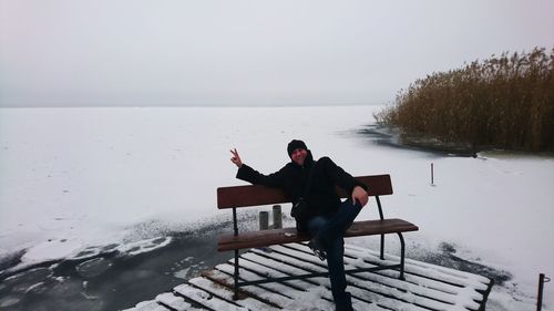 Man sitting on seat by lake against sky during winter