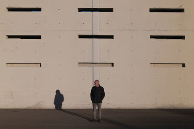 Full length of adult man in winter clothes standing against white wall with sunlight and shadow