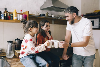 Smiling father toasting mugs with twin daughters sitting on kitchen counter at home