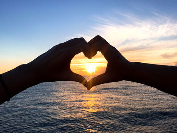 Cropped hands of couple making heart shape over sea against sky during sunset