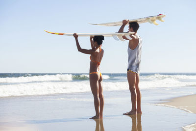 Couple carrying surfboard on head while standing at beach