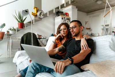 Concentrated young female in eyeglasses cuddling ethnic husband while watching movie together on laptop sitting on sofa at cozy home