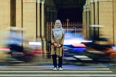 Full length portrait of smiling woman in hijab standing on city street with blur traffic in background at night