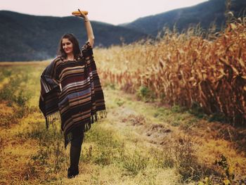 Portrait of smiling young woman with hand raised holding corn while walking on grassy field