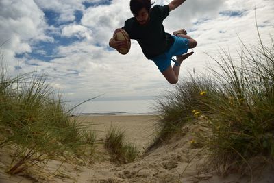 Man with american football jumping over grass at beach against sky