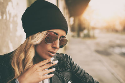 Fashionable young woman wearing jacket smoking cigarette outdoors