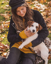 Smiling woman holding dog sitting at park during autumn