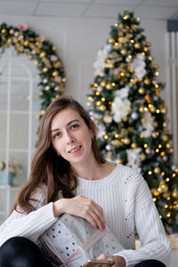 Girl in a white sweater sits with a christmas present on the background of a christmas tree.