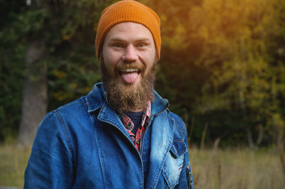 An attractive bearded man in a red hat and a denim jacket grimaces showing his tongue at the camera