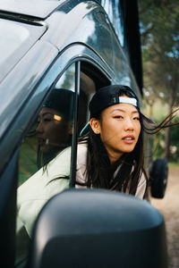 Happy young asian female traveler in cap looking out open window of camper van and enjoying freedom during trip through summer countryside