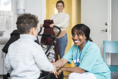 Smiling female nurse shaking hands with boy while mother standing in background at medical clinic