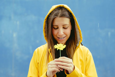 Smiling woman looking at yellow flower standing against blue background