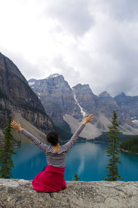 Rear view of woman with arms raised sitting on rock against mountains