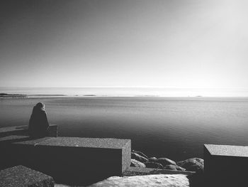 Rear view of woman sitting on retaining wall against sea during sunny day