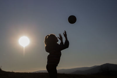 Silhouette little girl playing with ball in background against sky during sunset