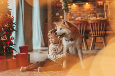 Candid authentic happy little boy in knitted beige sweater hugs dog with bow tie at home on xmas