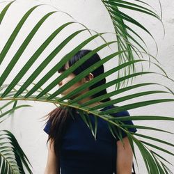 Close-up of young woman behind palm leaves against wall