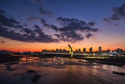 Illuminated city by the river against sky during sunset