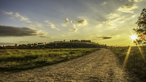 Dirt road amidst field against sky during sunset
