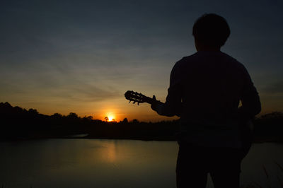 Rear view of silhouette man standing against lake during sunset