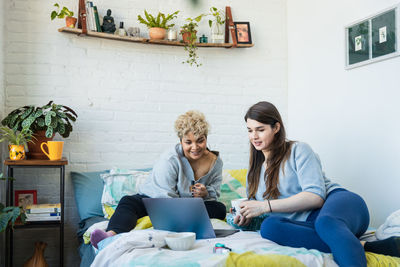 Woman showing laptop to roommate having coffee while sitting on bed at home