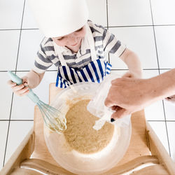 High angle cropped hand of woman pouring water while boy mixing batter on wooden ladder