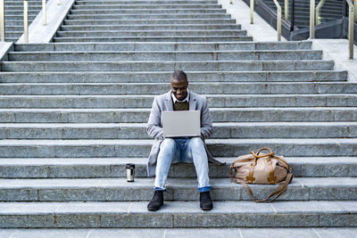 Smiling businessman with duffel bag and insulated drink container using laptop sitting on steps