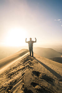 Full length of man standing with arms raised on sand dune against sky