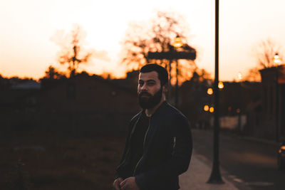 Portrait of young man standing in park against sky during sunset