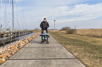 Grandfather pushing baby stroller with granddaughter on footpath against sky