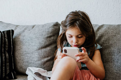 Adorable ethnic little girl in casual clothes and socks relaxing on comfortable sofa and playing video game with smartphone