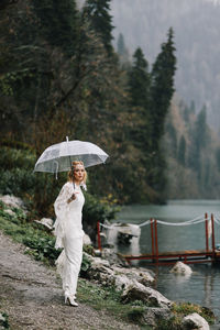 Beautiful young woman bride in a boho dress and with an umbrella stands in the rain in nature