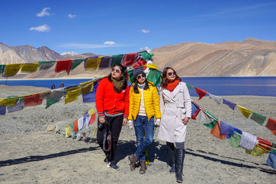 Full length of friends standing by colorful prayer flags against lake and mountains during sunny day