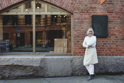 Full length of smiling baker standing on sidewalk against brick wall