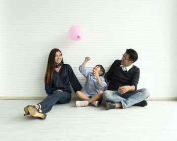 Smiling father and mother looking at daughter playing with balloon while sitting against wall