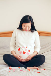 Smiling asian chinese pregnant woman sitting on bed. pregnant belly covered with red paper hearts